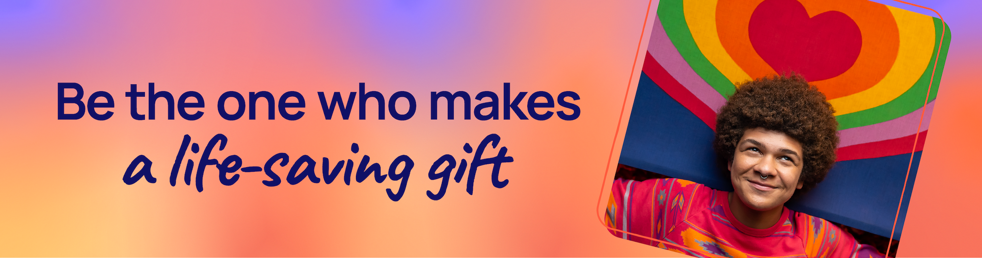 A photo of an LGBTQ+ young person in a colorful shirt, posing in front of a rainbox heart. Be the one to make a lifesaving gift.